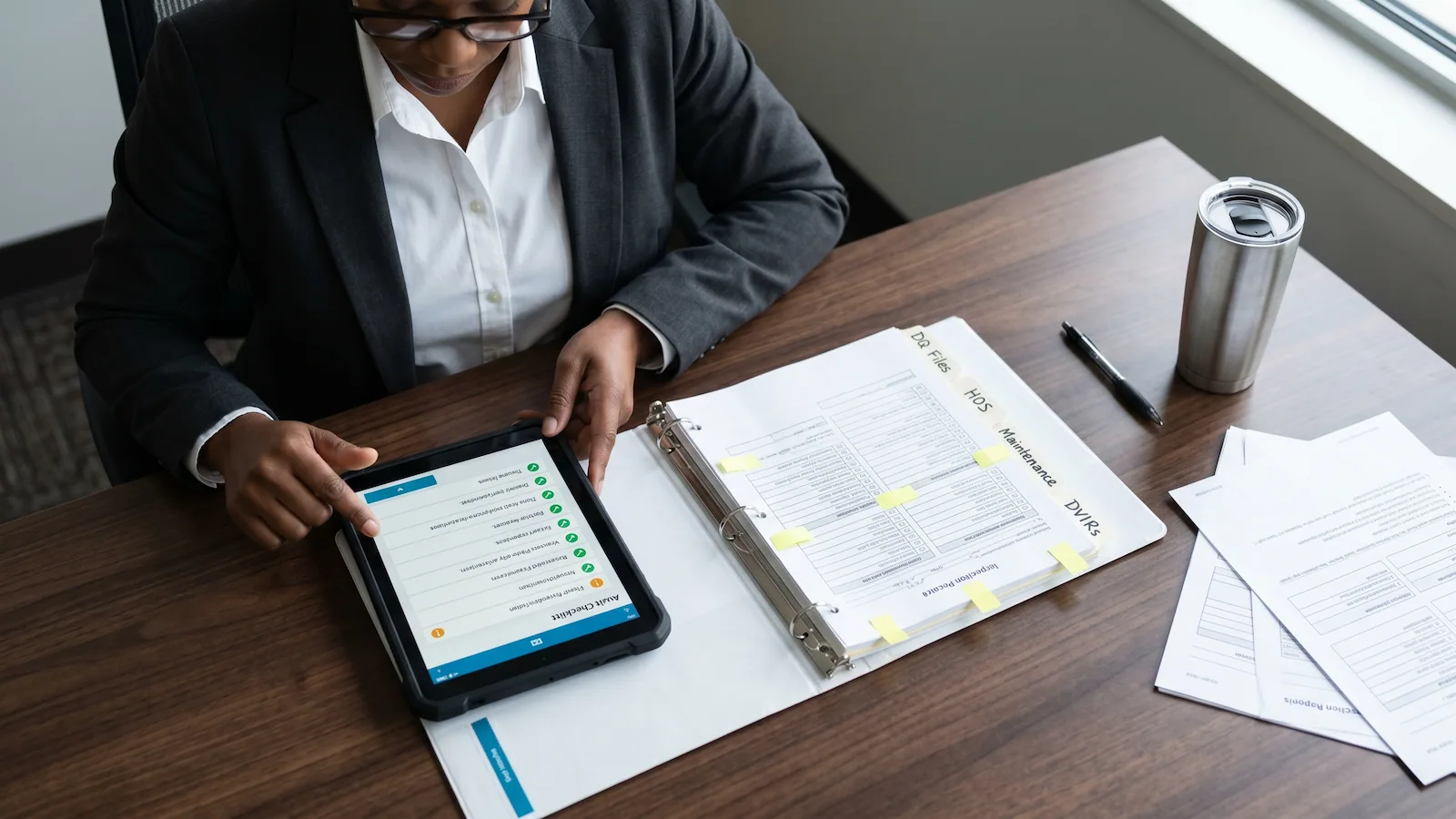 A fleet compliance officer at a desk reviewing a tablet checklist alongside a neatly organized binder of driver qualification and inspection records